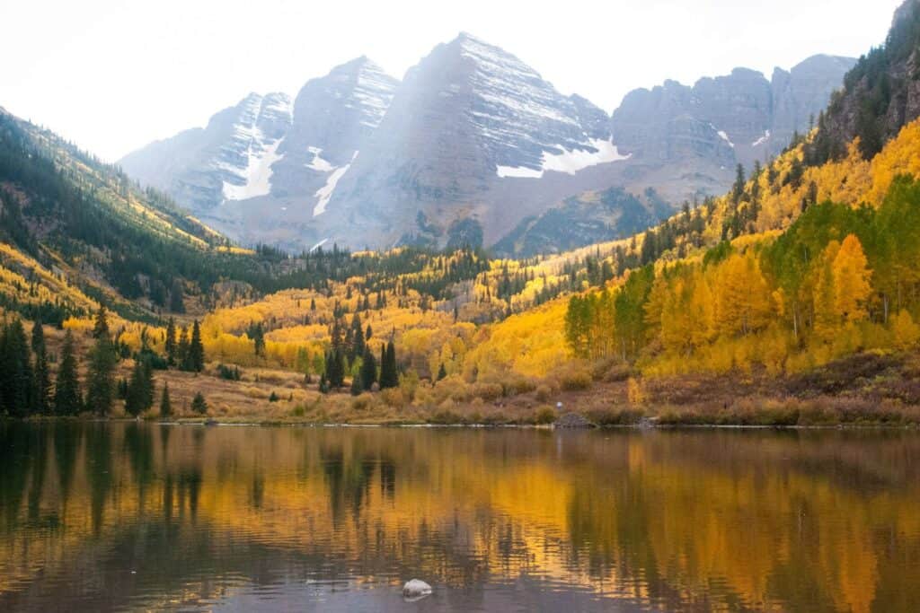 A serene lake reflecting the golden hues of autumn leaves against the backdrop of snow-dusted peaks, capturing the essence of a babymoon in Colorado