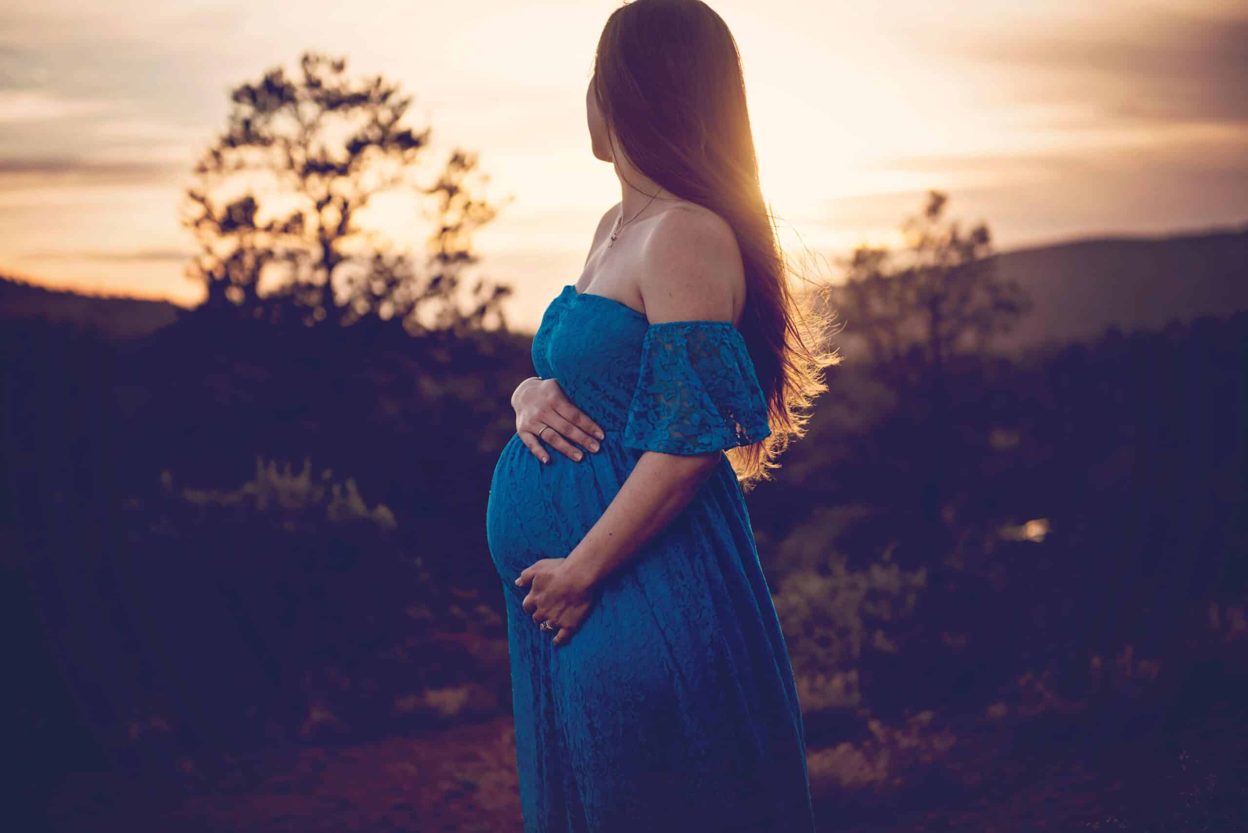 Silhouette of a pregnant woman in a flowing blue dress, standing peacefully in the desert near Palm Springs at sunset.