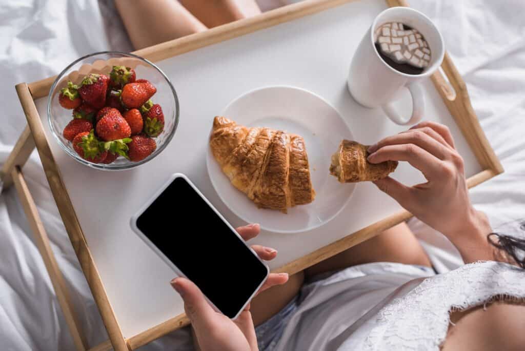 A woman eating a croissant looking at her phone while in bed