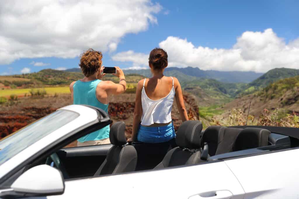 A couple pulled over in their white convertible taking a picture in Hawaii with their iphone, the perfect activity for their babymoon.
