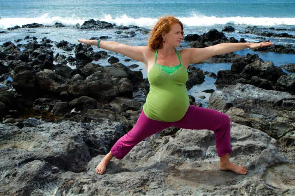 A red headed pregnant women doing warrior one yoga pose on a rocky beach in Hawaii on her babymoon